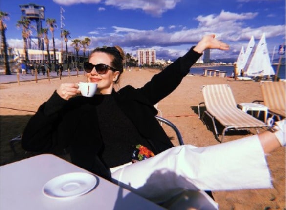 Cara wearing black jacket and white pant and holding a cup.